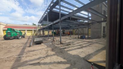 A construction site with steel framework for a new building, workers in safety gear, equipment, and dirt ground under a partly cloudy sky.