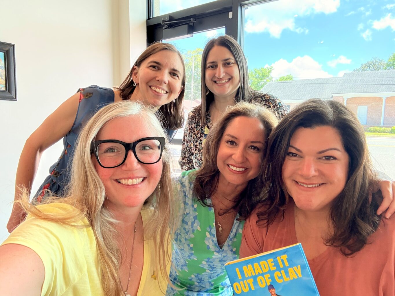 Five women smiling indoors near a window; one holds a book titled "I Made It Out Of Clay." Blue sky and buildings are visible outside.