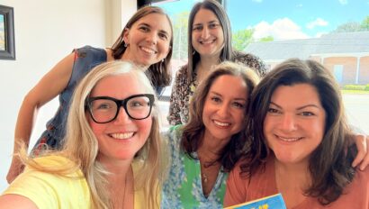 Five women smiling indoors near a window; one holds a book titled "I Made It Out Of Clay." Blue sky and buildings are visible outside.