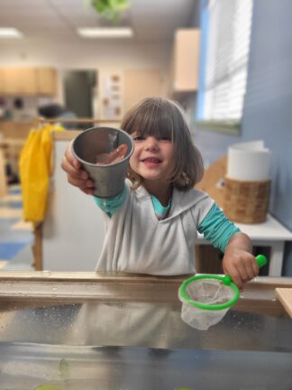 A young child smiling and holding a small metal cup with water and a toy fish, while standing at a water table and holding a green-handled net.