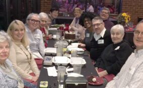 A group of people sit around a long table at a restaurant, smiling at the camera with food containers, drinks, and a chocolate cake in the center.