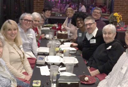 A group of people sit around a long table at a restaurant, smiling at the camera with food containers, drinks, and a chocolate cake in the center.