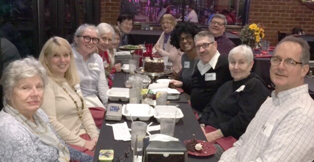 A group of people sit around a long table at a restaurant, smiling at the camera with food containers, drinks, and a chocolate cake in the center.