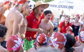 A swim coach in a straw hat and swimmers in red caps huddle and cheer together outdoors near event tents.