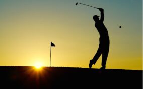 Silhouette of a golfer swinging a club at sunset, with a golf flag and the sun low on the horizon in the background.