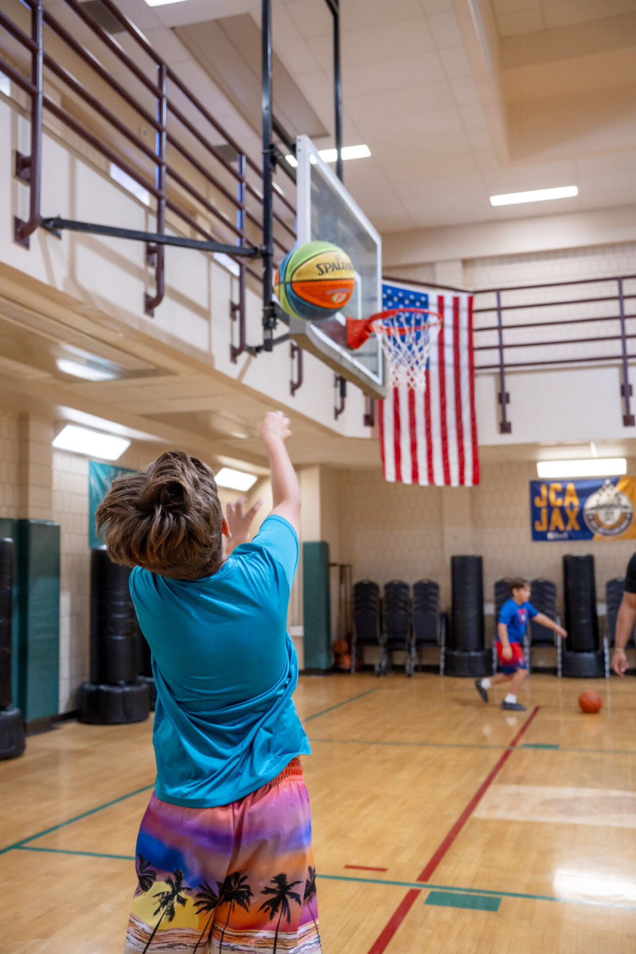 A boy in colorful shorts shoots a basketball toward a hoop in an indoor gym, with an American flag hanging in the background.
