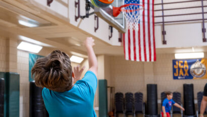 A boy in colorful shorts shoots a basketball toward a hoop in an indoor gym, with an American flag hanging in the background.
