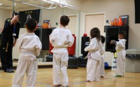 A martial arts instructor faces four young students in white uniforms and belts during a class in a studio with mirrored walls.