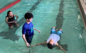 Two adults guide a child floating on their back in a swimming pool during a swim lesson, near a lane divider.