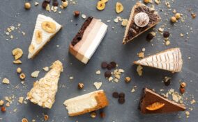 Six assorted slices of cake are arranged on a gray surface with scattered nuts and chocolate chips around them.