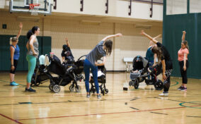 A group of women with strollers do side stretches together in a gymnasium.