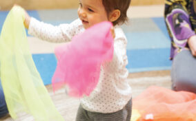 A toddler stands indoors, smiling and waving colorful scarves, with other scarves and adults visible in the background.