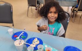 A young girl with curly hair sits at a table with craft supplies and a decorated cupcake, looking at the camera and smiling.