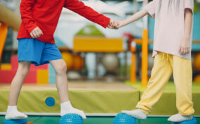 Two children holding hands while balancing on textured blue balance pods in a play area, wearing colorful clothes and socks.