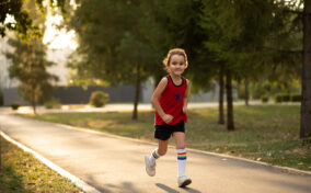 A young child runs along a paved path in a park surrounded by trees, wearing a red tank top, black shorts, white socks, and sneakers.