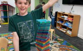 A smiling boy stands next to a tall, colorful tower made of magnetic tiles on a classroom table, raising one arm in excitement.