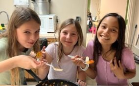 Three girls standing in a kitchen smile at the camera while tasting food from a pan on the stove.