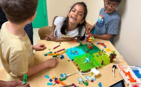 Four children gather around a table, building with LEGO bricks and smiling, with a laptop and LEGO pieces scattered on the tabletop.