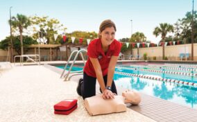 A woman kneels poolside practicing CPR on a training manikin, with a first aid kit nearby and a swimming pool in the background.