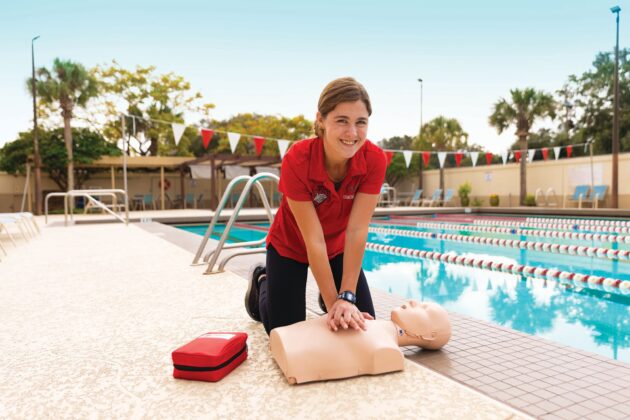 A woman kneels poolside practicing CPR on a training manikin, with a first aid kit nearby and a swimming pool in the background.