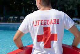 A person wearing a white "LIFEGUARD" shirt with a red cross sits by a swimming pool, facing the water.