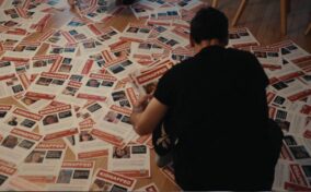 A person sits on a wooden floor covered with numerous missing person flyers labeled "KIDNAPPED," each showing photos and details of different individuals.