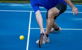 A person wearing a blue shirt and black shorts reaches down with a paddle to hit a yellow pickleball on an outdoor blue court.