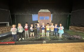 A group of young children stand in a row on a stage, each holding a craft project, with a painted backdrop resembling a house behind them.