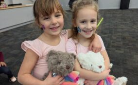 Two young girls wearing light pink dresses and face paint smile while holding teddy bears dressed in rainbow outfits in a carpeted indoor space.