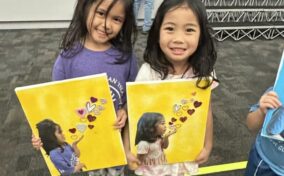 Two young girls smiling and holding colorful art prints featuring themselves blowing heart-shaped bubbles on a yellow background.