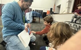 An adult kneels on the floor showing a book to two children sitting in front of him, while another child sits on the stage in the background.