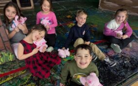 Six young children sit on a colorful painted floor, smiling and holding small pink stuffed animals.