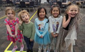 Five young children stand side by side indoors, smiling at the camera. They are dressed in colorful outfits, and several empty chairs and tables are visible in the background.