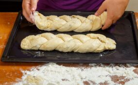 Two braided loaves of raw bread dough are placed on a baking sheet, with hands adjusting one loaf. The table and tray are dusted with flour.