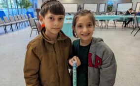 Two children stand indoors, the boy wears a brown jacket and the girl wears a gray jacket over a green top. They both face the camera and smile, with empty chairs and tables in the background.