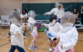 Four children in fencing gear practice with foils in a gymnasium, while several people watch from the bleachers in the background.