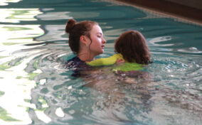 An instructor in a pool supports a young child wearing a green swim shirt as they practice swimming together.