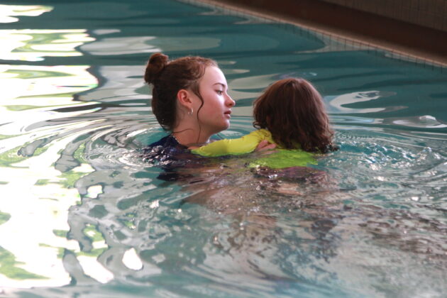 An instructor in a pool supports a young child wearing a green swim shirt as they practice swimming together.
