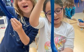 Two young girls in a classroom stretch blue slime, smiling and looking at the camera. Other children and backpacks are visible in the background.