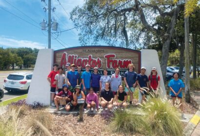 A group of people poses in front of the St. Augustine Alligator Farm Zoological Park entrance sign on a sunny day.