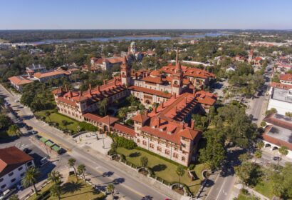 Aerial view of a large historic building with red-tiled roofs, surrounded by roads and greenery in an urban area under a clear sky.