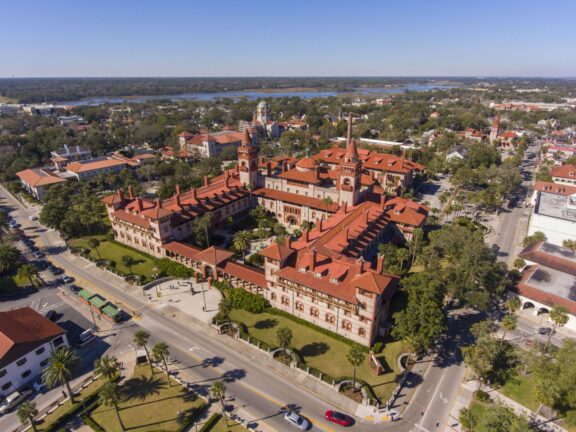 Aerial view of a large historic building with red-tiled roofs, surrounded by roads and greenery in an urban area under a clear sky.