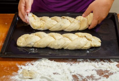 A person places two braided dough loaves on a black baking tray, with flour scattered on the wooden surface below.