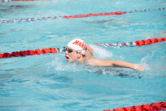 A swimmer wearing goggles and a white cap labeled "MARA" swims the butterfly stroke in a pool, separated by orange and white lane dividers.