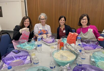Four women sit at a table with baking supplies, bowls, flour, and bottled water, smiling at the camera in a brightly lit room.