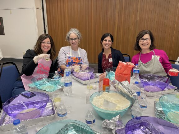 Four women sit at a table with baking supplies, bowls, flour, and bottled water, smiling at the camera in a brightly lit room.
