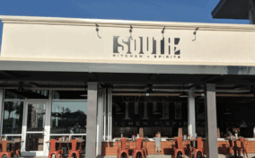 Outdoor view of South Kitchen + Spirits restaurant, featuring a white facade, large windows, and red chairs at tables on the patio.