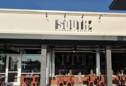 Outdoor view of South Kitchen + Spirits restaurant, featuring a white facade, large windows, and red chairs at tables on the patio.