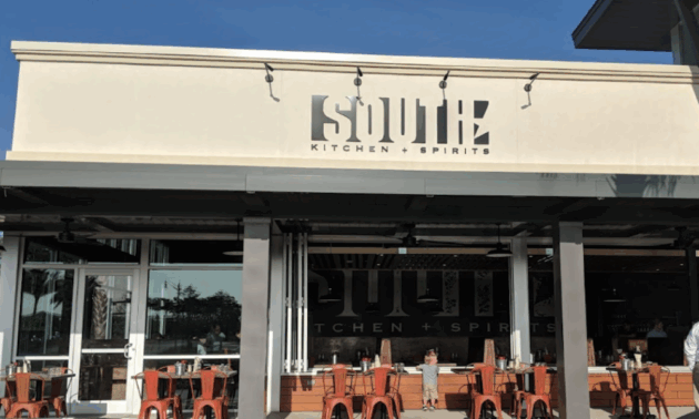 Outdoor view of South Kitchen + Spirits restaurant, featuring a white facade, large windows, and red chairs at tables on the patio.