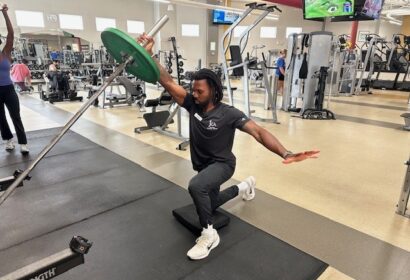 A man performs a half-kneeling landmine press with a weight plate in a gym, balancing on one knee and extending his opposite arm for stability.